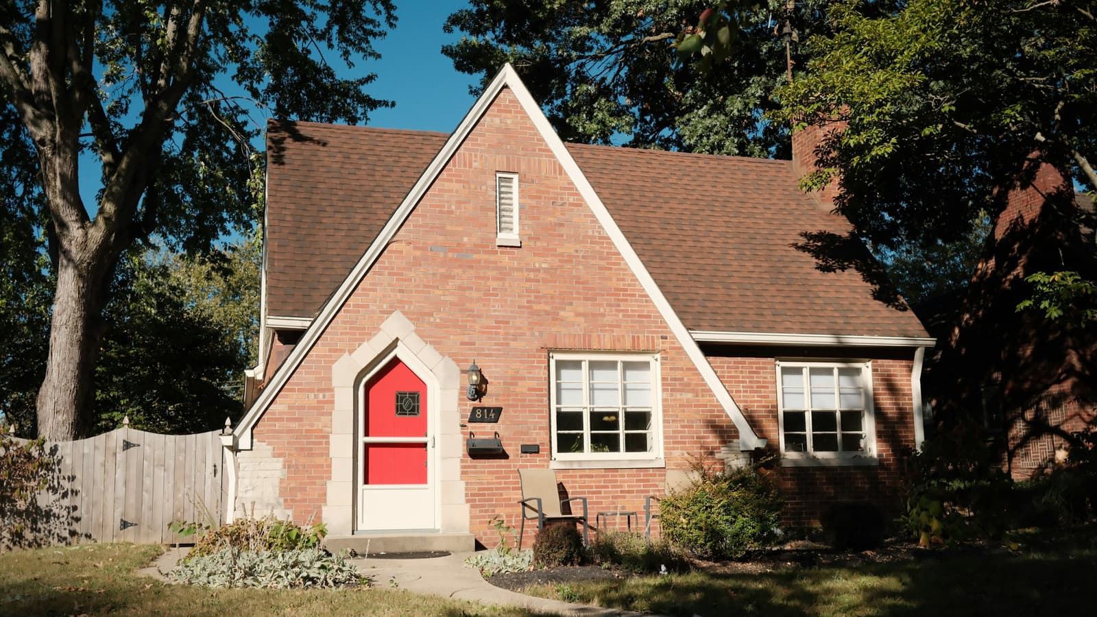 Red brick Tudor-style cottage with a steep brown asphalt-shingle roof, red arched front door and mature trees